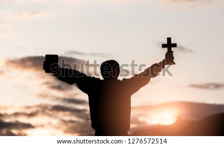 Young christian holding cross and bible with light sunset background, christian silhouette concept.