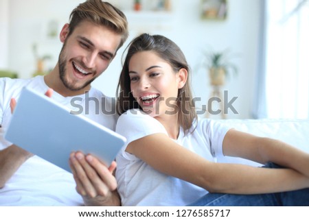 Young couple watching media content online in a tablet sitting on a sofa in the living room. Royalty-Free Stock Photo #1276585717