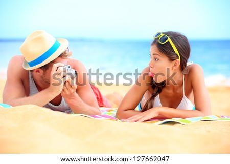 Young couple having fun on beach with vintage retro camera. Funky young interracial couple playful on beach during summer holidays travel vacation. Caucasian man and Asian girl making funny face.