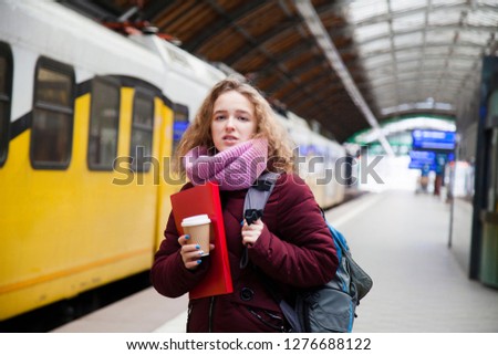 A young girl with curly hair and a backpack is standing near the train at the train station. Holding a paper cup of coffee and a red folder for papers Royalty-Free Stock Photo #1276688122
