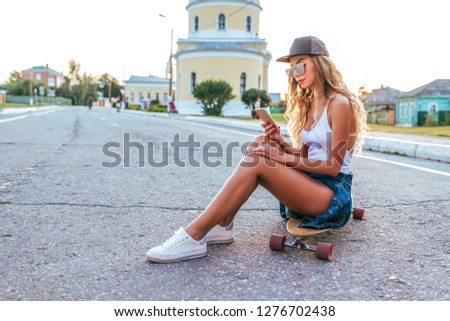 A girl in white bodysuit, sits skateboard longboard, long hair, tanned skin, in sunglasses. In hands of phone holding, communicates on Internet. Concept communication of friends in social networks.