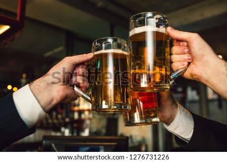 Picture of three mugs of beer in men hands. People wear suits. They are in bar.