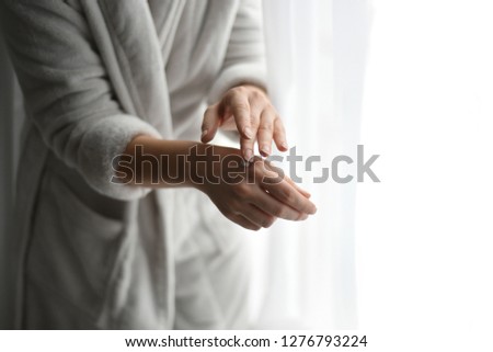 Young bride preparing for her big day. Applying a light moisturizing cream on her hands. Wearing soft, gentle, fluffy and light grey bathrobe. Standing in front of the window. Banner free copy space.