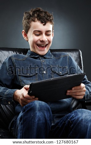 Joyful young boy sitting on leather chair, playing on tablet. Gray background