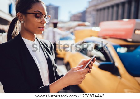 Attractive afro american girl in optical spectacles tracking yellow cab via useful application on smartphone connected to 4G, confident businesswoman checking notification on modern cellphone
