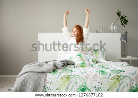 Smiling redhead girl waking up in big comfortable bed with floral bedding and grey blanket, real photo of grey bedroom design Royalty-Free Stock Photo #1276877182