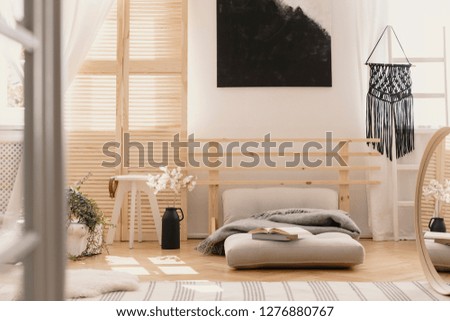 Black handmade macrame on white wooden ladder in bright bedroom interior with wooden furniture and beige futon with grey warm blanket, real photo with mockup on the empty wall