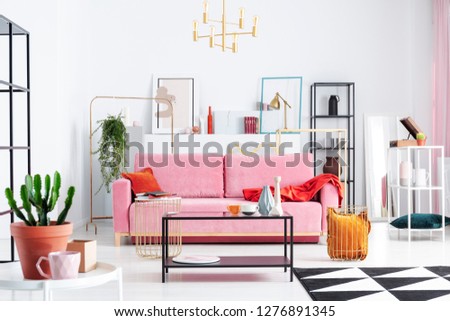 Patterned carpet and pink couch in white apartment interior with posters and lamp above table. Real photo