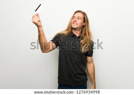 Blond man with long hair over white wall holding a credit card and thinking