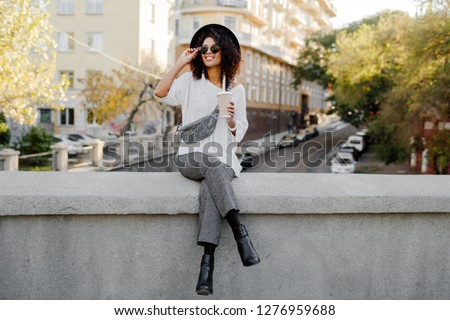 Full height image Playful black woman with Afro hairs sitting on the bridge and having fun. Wearing leather boots and whoop  trendy trousers . Travel mood. Happy leisure time in old European city. 