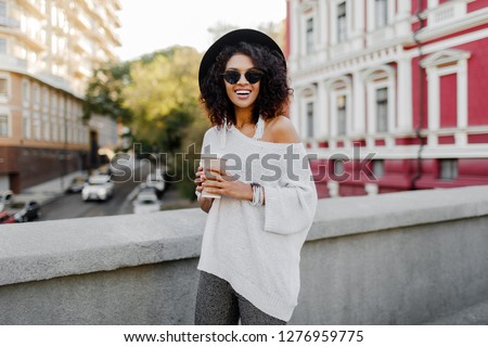 Soft toned lifestyle outdoor image of blissful black woman walking in spring  city with cup of cappuccino or hot tea. Hipster outfit. Oversize white sweater, black hat, stylish accessories. 