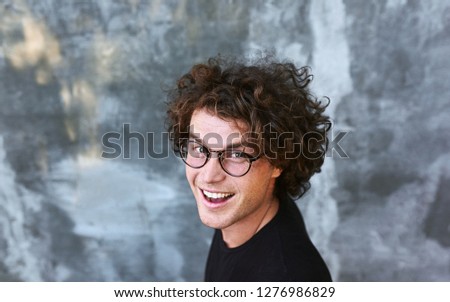 Top view portrait of handsome happy smiling male with curly hair, wears spectacles posing for social advertisement, isolated on gray concrete wall with copy space for your promotional information