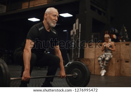 People, age, sports, vitality and powerlifting. Picture of muscular fit mature retired man with strong arms exercising with barbell posing in gym with his young female instructor in background