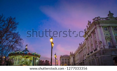 A vintage carousel is located next to the Royal Palace at Plaza Oriente square in christmas with the colorful pink sky at blue hour, sunset in the background