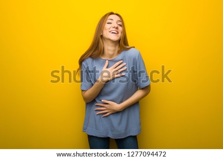 Young redhead girl over yellow wall background smiling a lot while putting hands on chest