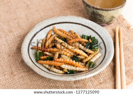 Food Insects: Bamboo worm (Bamboo Caterpillar) insect fried crispy for eating as food items in plate with chopsticks and tea on sackcloth, it is good source of protein edible for future food concept. Royalty-Free Stock Photo #1277125090