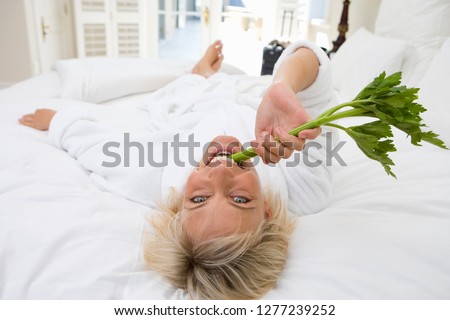 Woman wearing white bathrobe in hotel room biting on celery at camera