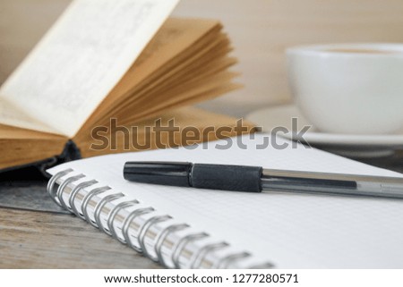 The pen is on a notebook, in the background is a cup and an open book.