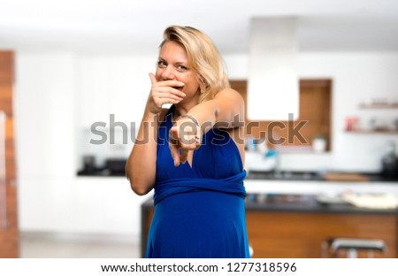 Pregnant blonde woman with blue dress pointing with finger at someone and laughing a lot in her house