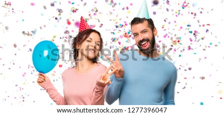couple with balloons and birthday hats smiling and showing victory sign with both hands with confetti in a party