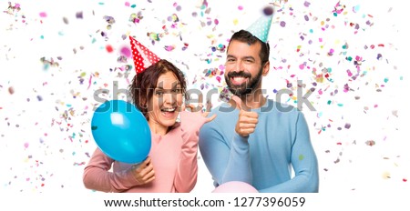 couple with balloons and birthday hats showing ok sign and giving a thumb up gesture with the other hand with confetti in a party