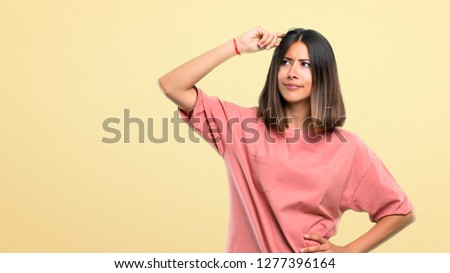 Young girl with pink shirt having doubts and with confuse face expression while scratching head on yellow background