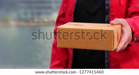 Adult hand holding brown box with red shirt, parcel shipping delivery business for digital online shopping marketing idea or post service concept on blurred background in daylight with Copy space
