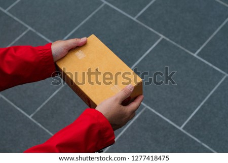 Adult hand holding brown box with red shirt, parcel shipping delivery business for digital online shopping marketing idea or post service concept on blurred background in daylight with Copy space.