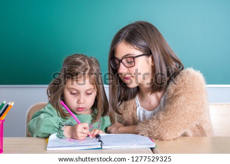 older sister looks at her younger sister as she draws in a notebook.