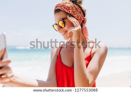 Magnificent european woman taking picture of herself on sea background. Good-looking tanned girl in glasses making selfie with phone.