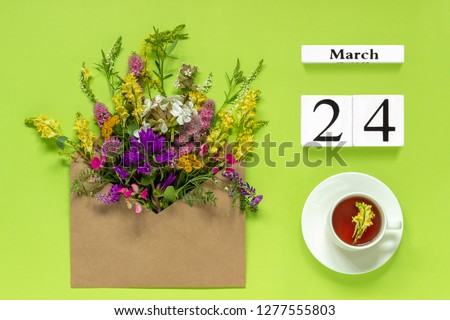 Wooden cubes calendar March 24. Cup of herbs tea, kraft envelope with multi colored flowers on green background. Concept hello spring Creative Top view Flat lay.