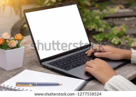 Female hands typing on laptop computer keyboard with blank white screen, notebook and pen on wooden table at green garden background. Online education concept.