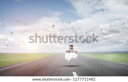 Young little boy keeping eyes closed and looking concentrated while meditating on cloud above the road with beautiful and breathtaking landscape on background.