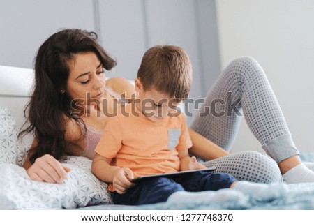Little kid watching cartoons on tablet while sitting on the bed while his mother watching cartoons with him. Bedroom interior.