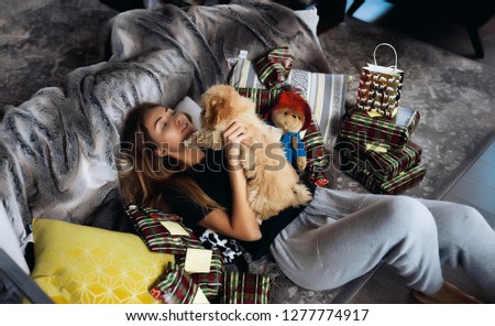 Winter picture of pretty blonde woman in black shirt and grey pants. Lying on a couch with little cute puppy in hands. A lot of presents around, Christmas holidays. Warm cozy atmosphere of happiness