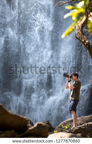 Asian man traveler and photographer using digital camera taking photo at waterfall. Travel lifestyle or nature photography concepts