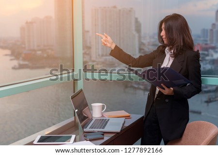 Asian woman holding folder of documents, pointing up on copy space and standing in front of windows in an office building overlooking the city and river. 