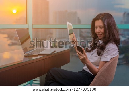 Asian businesswoman having business call in office, her workplace, writing down some information. Woman using Laptop, Tablet and looking at smartphone screen. 
