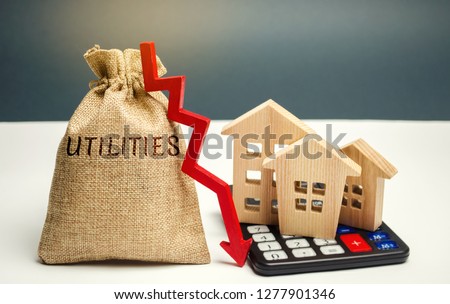 Money bag with the word Utilities and an arrow down and wooden houses on the calculator. Reduced prices for utilities. Low prices for electricity, heating. Payment for the use of water. Royalty-Free Stock Photo #1277901346