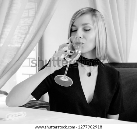 Glamour young blonde sitting in restaurant holding wine glass in dress. Orientation of the picture is square. Square pattern. Black and white photo. 