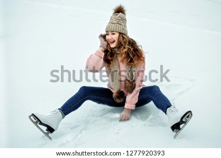 Beautiful girl in warm clothes sitting on ice rink after falling and laughing