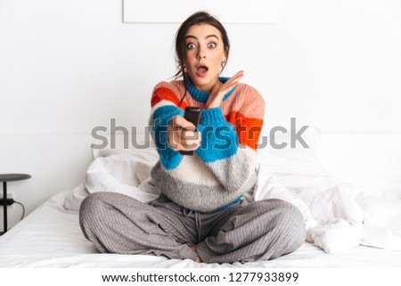 Photo of excited woman 30s holding remote control while sitting in bed at home