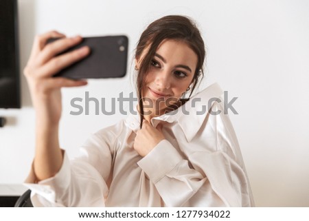 Photo of seductive woman 20s wearing silk clothing taking selfie photo on black smartphone while standing in kitchen in the morning