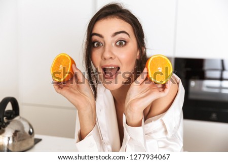 Photo of positive woman 30s wearing silk clothing holding two pieces of orange during breakfast in kitchen at home
