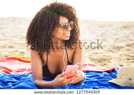 african origin girl relaxing on beach. afro hair woman wearing sunglasses lying on beach using phone listening to trendy  music with ear phones.