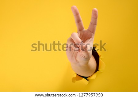 Hand showing peace gesture through torn yellow paper background. Victory, V sign. Royalty-Free Stock Photo #1277957950