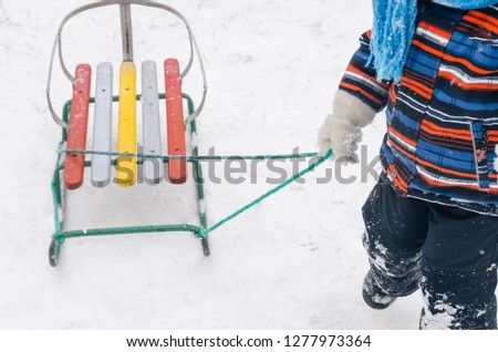 Winter season, snowing outside. A little boy in colored coat with sledge in his hand is running on top of the snowy hill. Riding the sled concept.