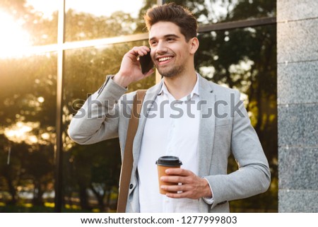 Photo of young businessman in suit talking on mobile phone while standing outdoor near building with takeaway coffee