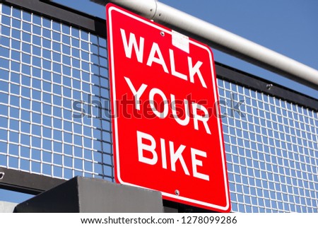Red sign walk your bike hanging on wire fence with blue sky as background