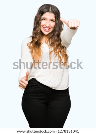 Young beautiful woman wearing white sweater approving doing positive gesture with hand, thumbs up smiling and happy for success. Looking at the camera, winner gesture.